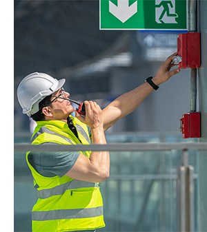 A person wearing a hardhat and hi-viz jacket in an industrial workplace presses the fire alarm under an emergency exit safety sign.