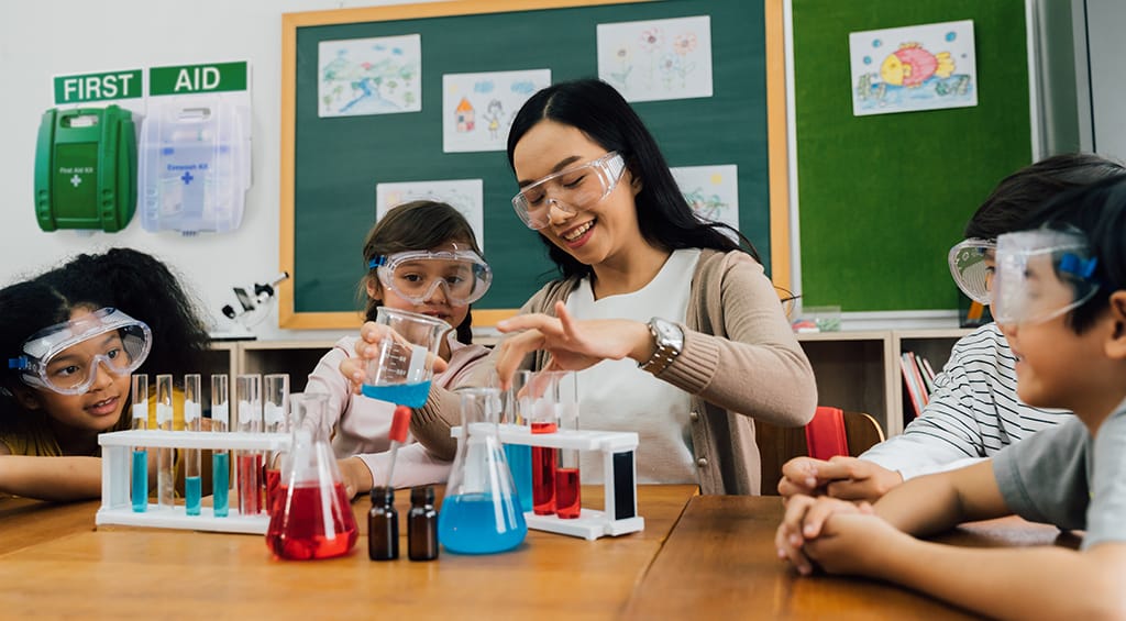 Teacher instructs young students in a science class, wearing protective gear with a first aid kit on the wall