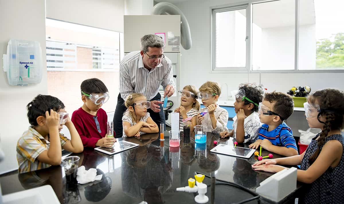 Science teacher performs experiment with primary school class, all wearing safety goggles with eye wash first aid kit on the wall