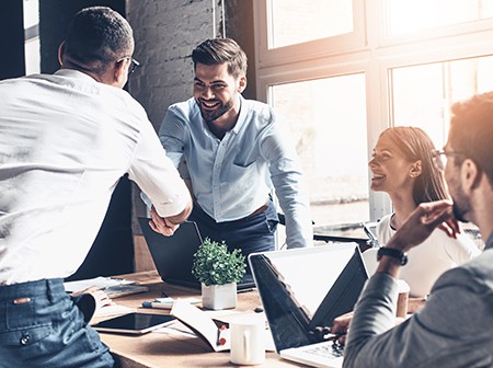 Group of people in workplace meeting room spread viruses by shaking hands and sitting close around a table with devices and mugs.