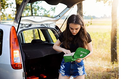 A car on the side of a sunny rural road, with the boot open and a woman looking through a first aid kit for first aid supplies