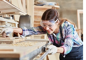 	 Woman wears safety goggles to protect her eyes from sawdust particles, as she uses machinery to cut wood