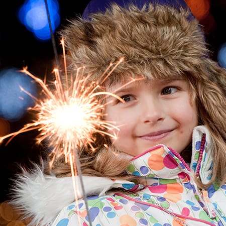 Young girl in bright, spotted coat and blue, fur-rimmed hat smiles holding sparkler