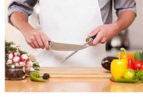 Person uses knife safety skills to sharpen knife carefully on kitchen board, with vegetables to the side