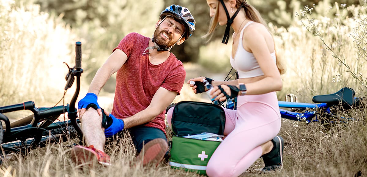 On a forest path, one cyclist administers first aid to another cyclist using a comprehensive sports first aid kit; bikes lie nearby 