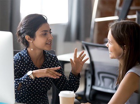 Two women in office talking animatedly over coffee and smiling, showing reduced stress levels from communication