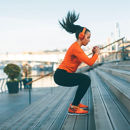 Woman outside in city exercising by jumping up steps next to lake. In squat position, with hair flying and hands clenched in front