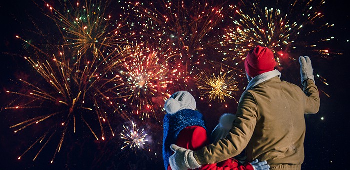 a family of two adults and one child stand close together, in winter clothing, watching a fireworks display at night