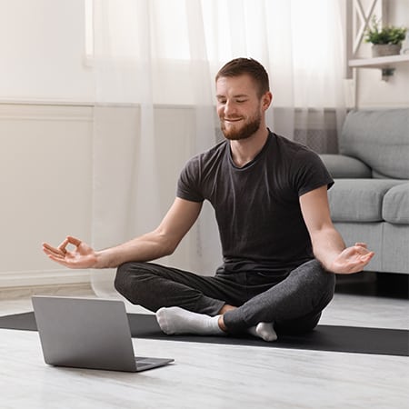 Smiling man sat cross-legged on yoga mat in front of laptop, hands resting on knees, facing up with thumb and forefinger touching
